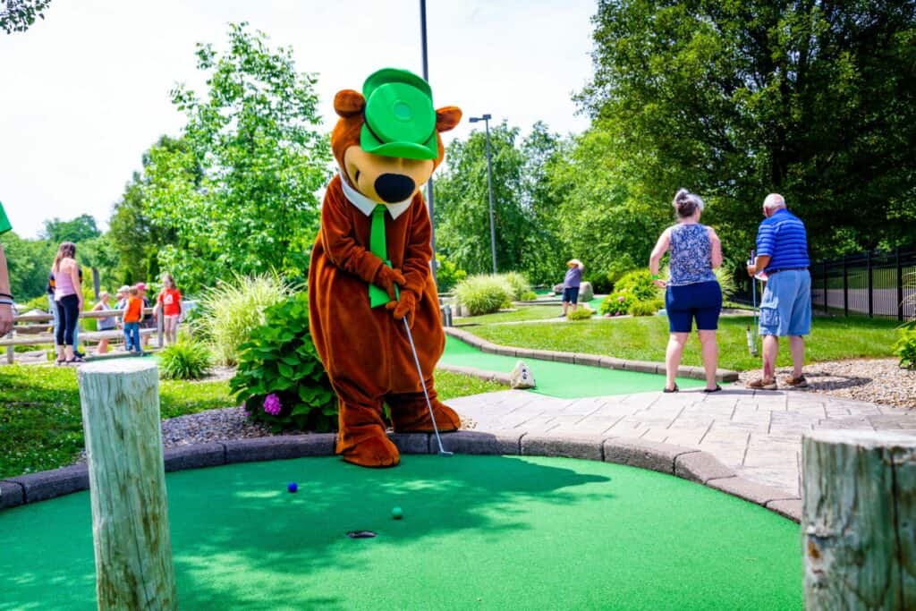 A mascot in a green hat is playing mini golf.