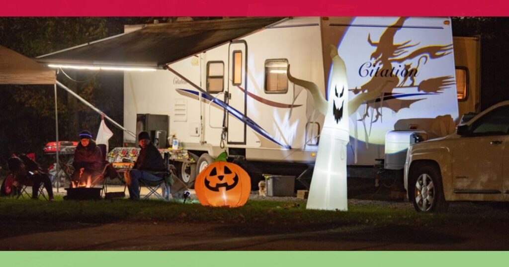 Two people sit by a campfire near an RV decorated for Halloween with a lit jack-o'-lantern, a ghost decoration, and ghost projection on the RV exterior.