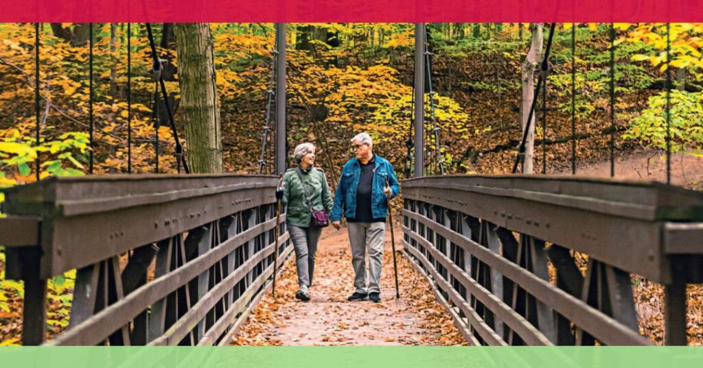 Two older adults walk together on a wooden bridge in a forest with autumn leaves, surrounded by trees with yellow and orange foliage.