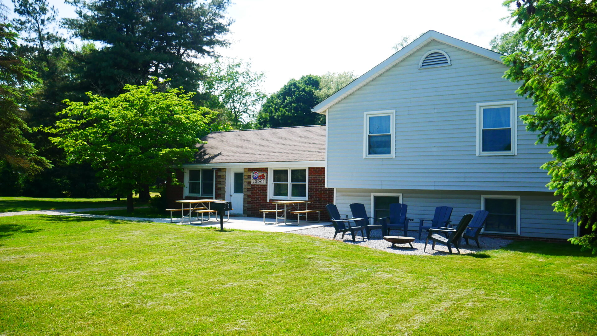 A light gray house with a patio featuring picnic tables and a seating area with Adirondack chairs, reminiscent of cozy cabins. The house is surrounded by green lawn and trees.