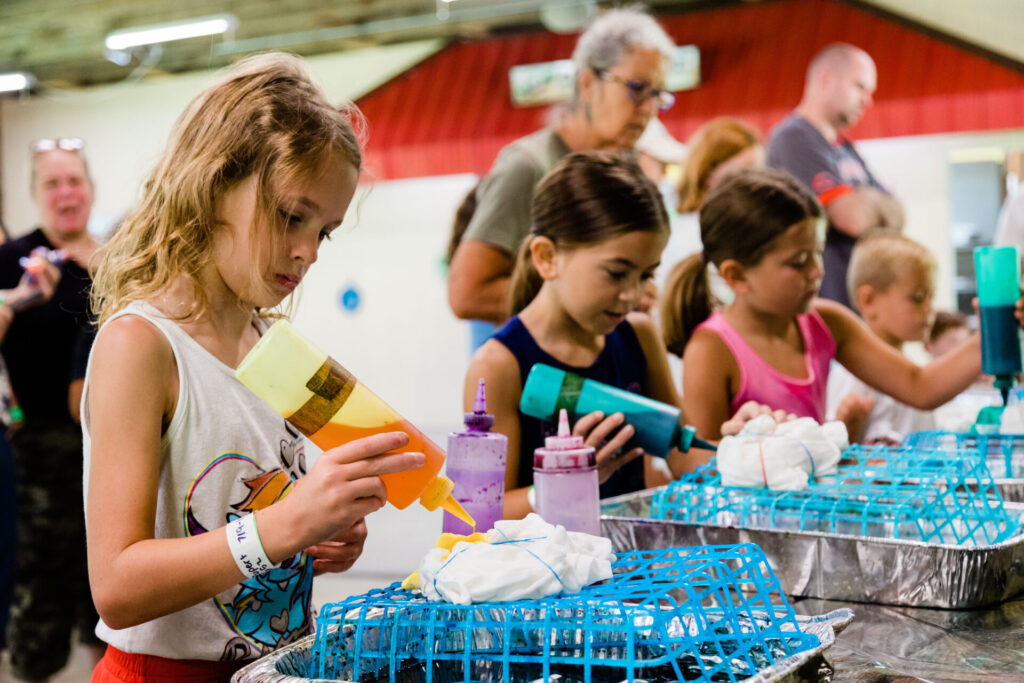 Children focused on tie-dyeing shirts at a table with various colored bottles, as adults supervised them in a busy indoor setting that buzzed with creative job opportunities.