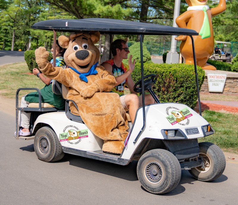 Boo Boo bear waves energetically while sitting on a moving golf cart rental with two others, enjoying a sunny day at the park.