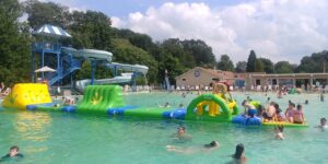 People swim and play on an inflatable obstacle course in an outdoor pool with water slides and trees in the background. People swim and play on an inflatable obstacle course in an outdoor pool with water slides and trees in the background.