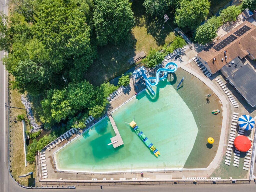 Aerial view of an outdoor swimming pool with a curved blue waterslide, floating obstacle course, lounge chairs, and surrounding trees.