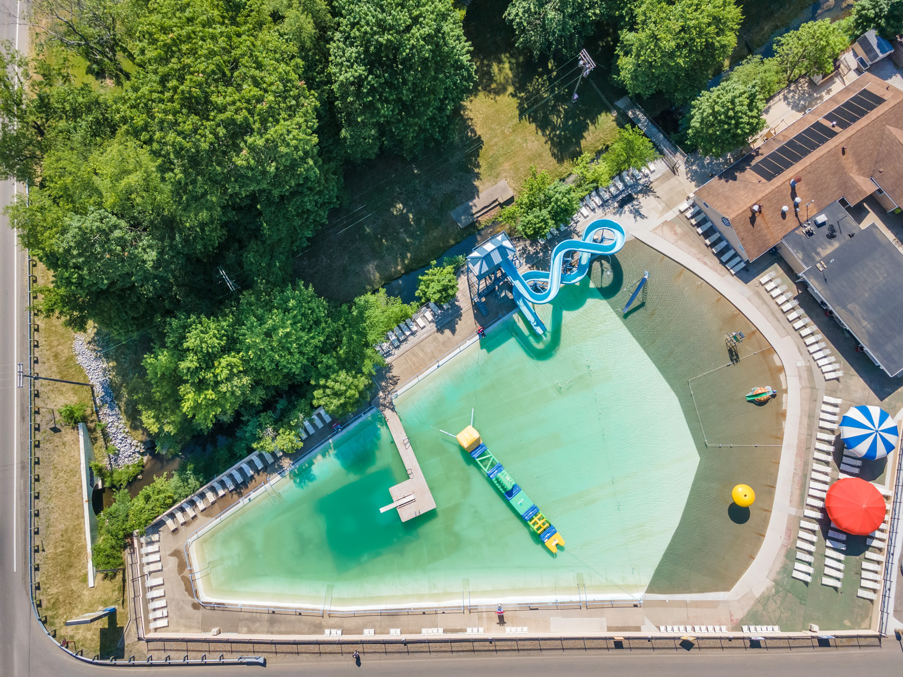 Aerial view of an outdoor swimming pool with a curved blue waterslide, floating obstacle course, lounge chairs, and surrounding trees.