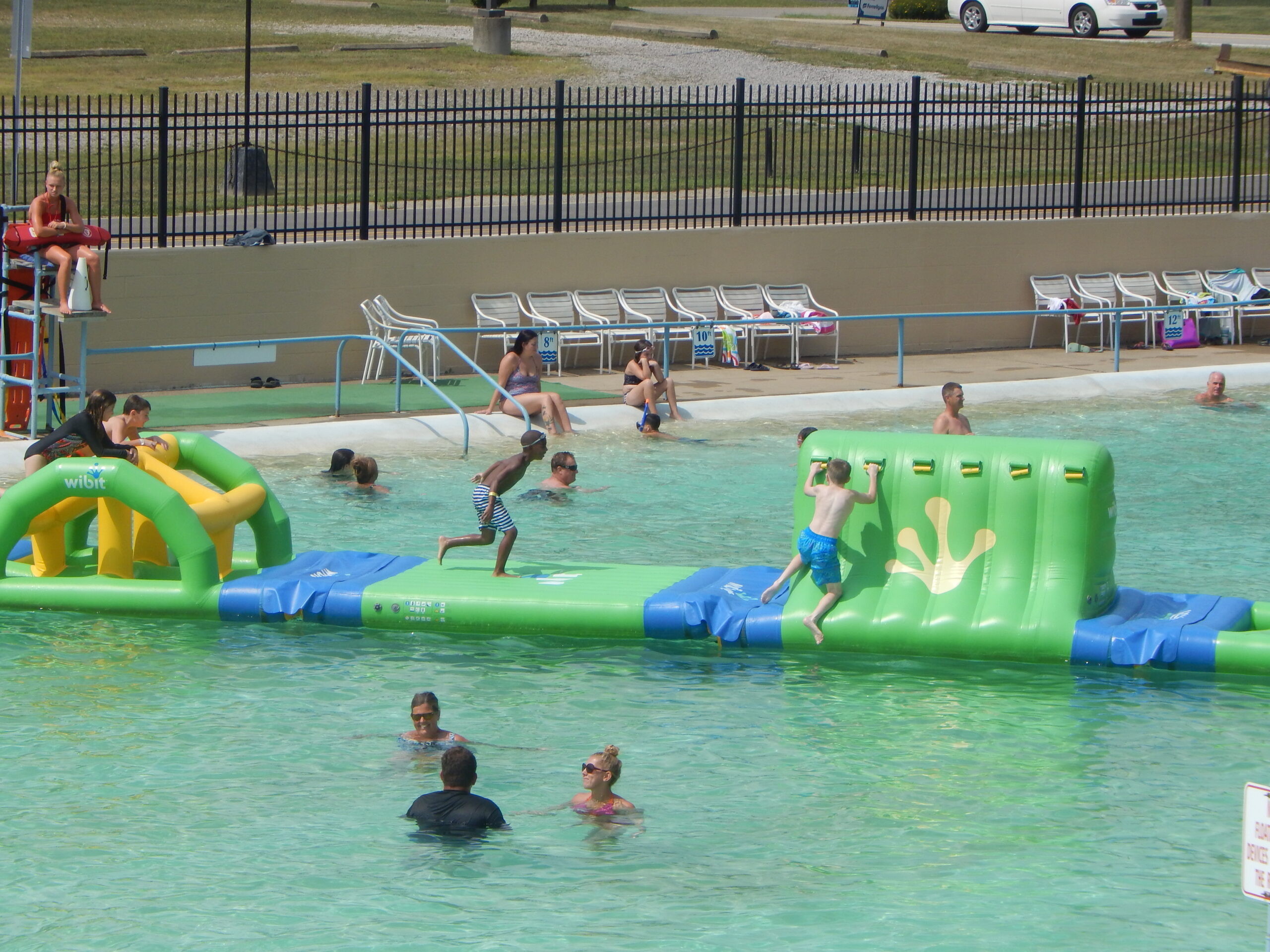 Children play on an inflatable obstacle course in a public outdoor pool while adults and other children swim nearby. A lifeguard watches from a stand.
