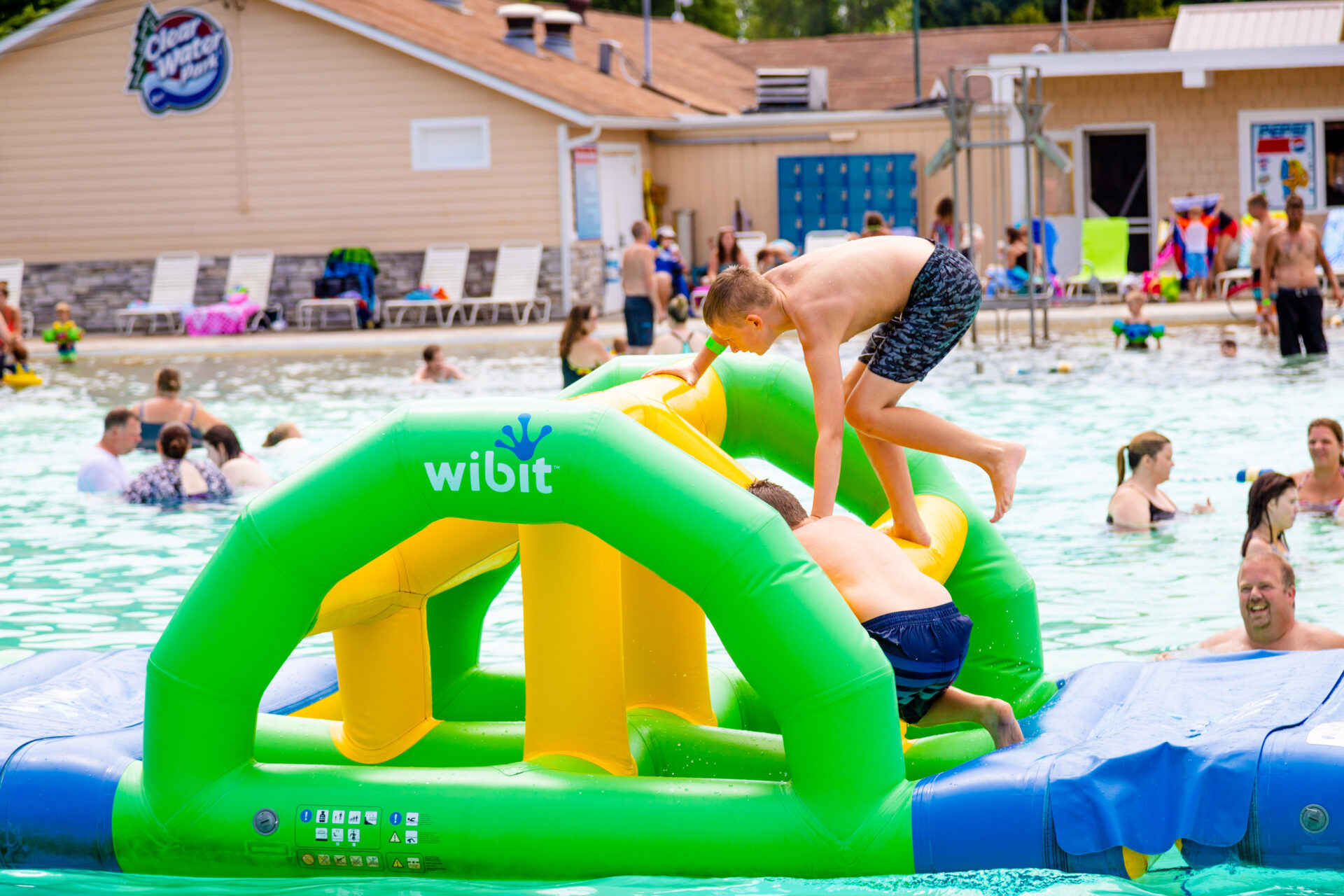 Children climb on a green and yellow inflatable obstacle in a crowded outdoor public pool with people swimming and lounging nearby.