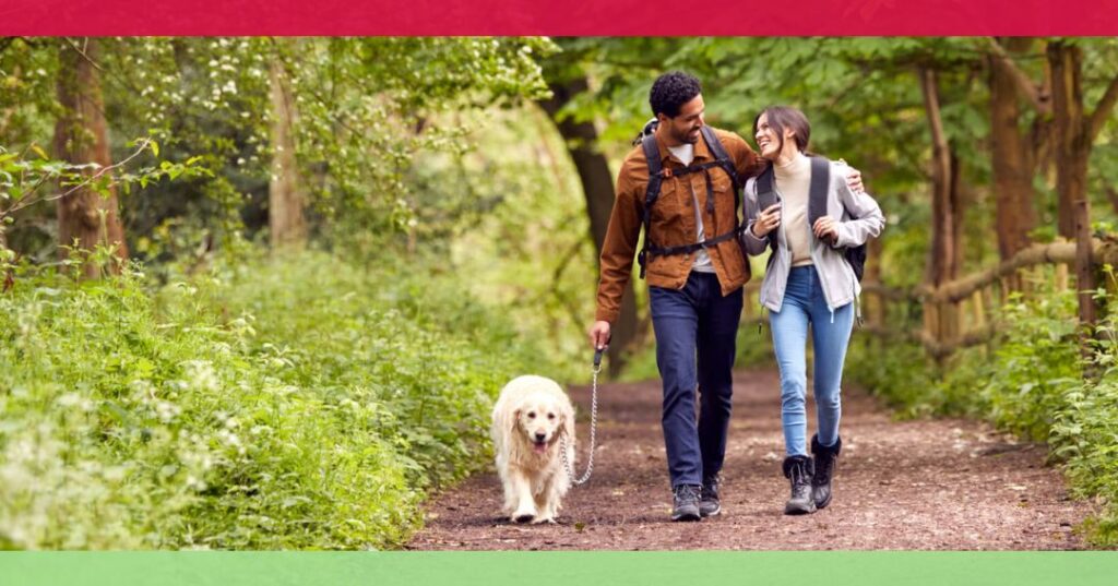 A man and woman walk a dog together on a wooded path, surrounded by greenery. The man has his arm around the woman, and both are smiling.