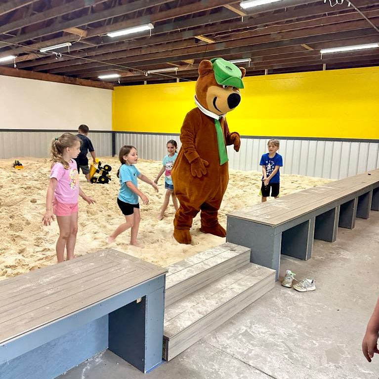 A person in a bear costume stands on a platform in an indoor sandbox area while children play around in the sand.