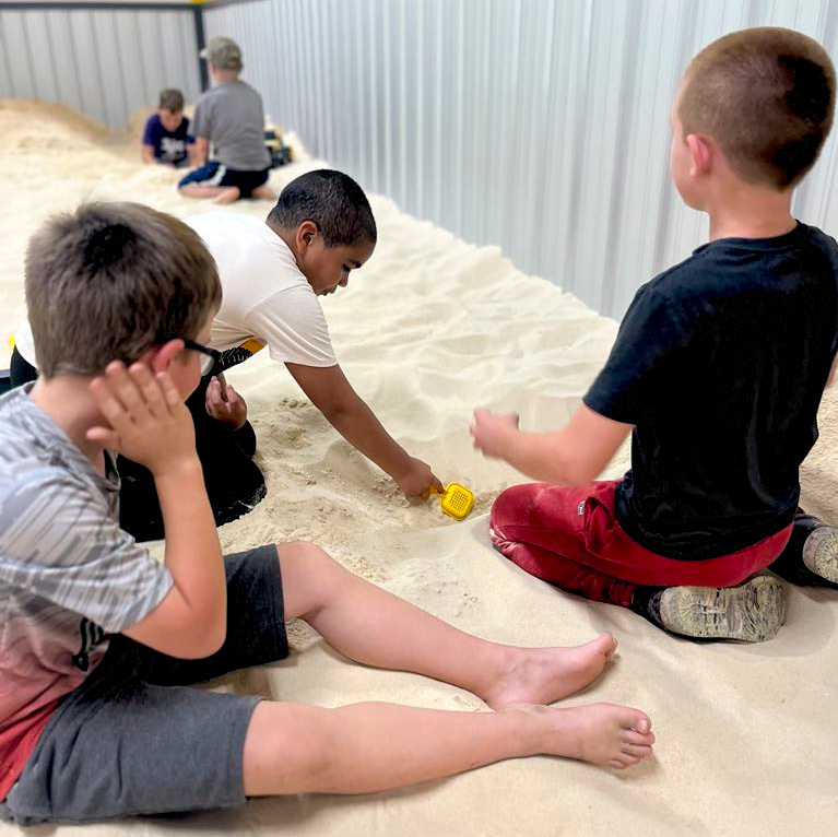 Three boys sit and play in an indoor sandbox with shovels, while two more children are visible in the background against a metal wall.