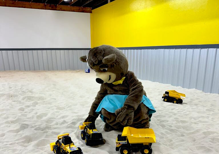 Person in a bear costume sits on sand, playing with yellow toy dump trucks. A blue plastic bucket lies nearby. The room has yellow and grey walls.