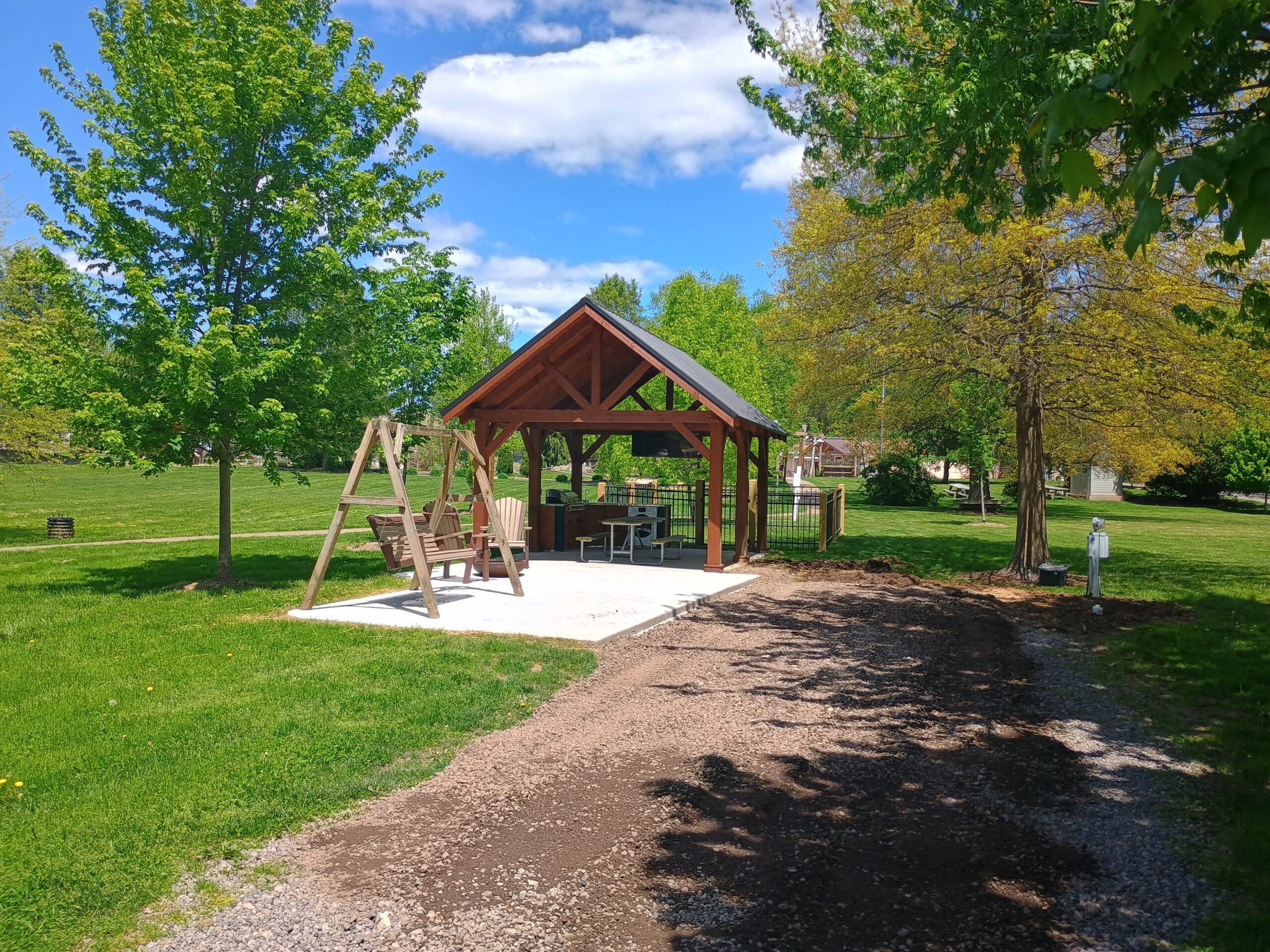 A wooden pavilion with a picnic table and swing sits on a concrete slab in a grassy park surrounded by trees under a partly cloudy sky.