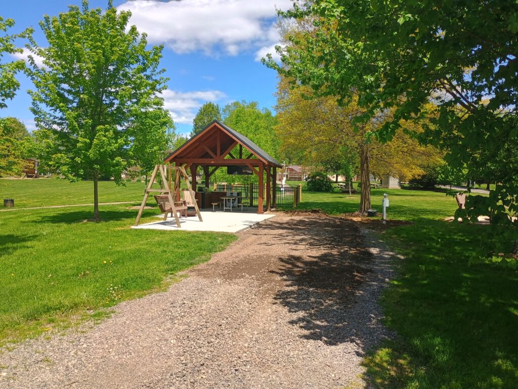 A wooden pavilion with benches stands on a concrete pad in a grassy park, surrounded by green trees under a partly cloudy sky. A gravel path leads to the pavilion.