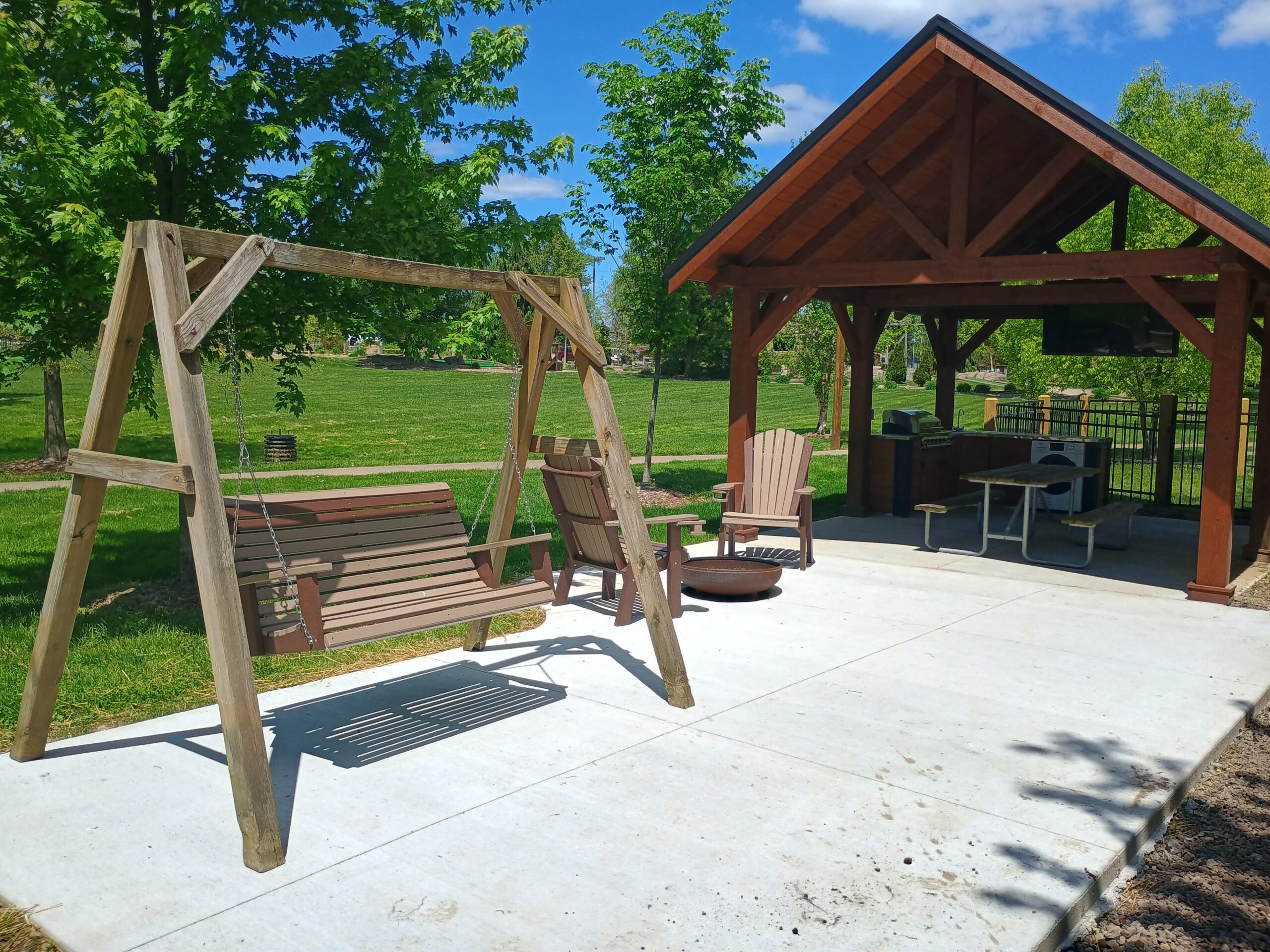 A wooden swing and Adirondack chairs are set on a concrete patio beside a covered picnic shelter with a table and outdoor grill in a park setting.