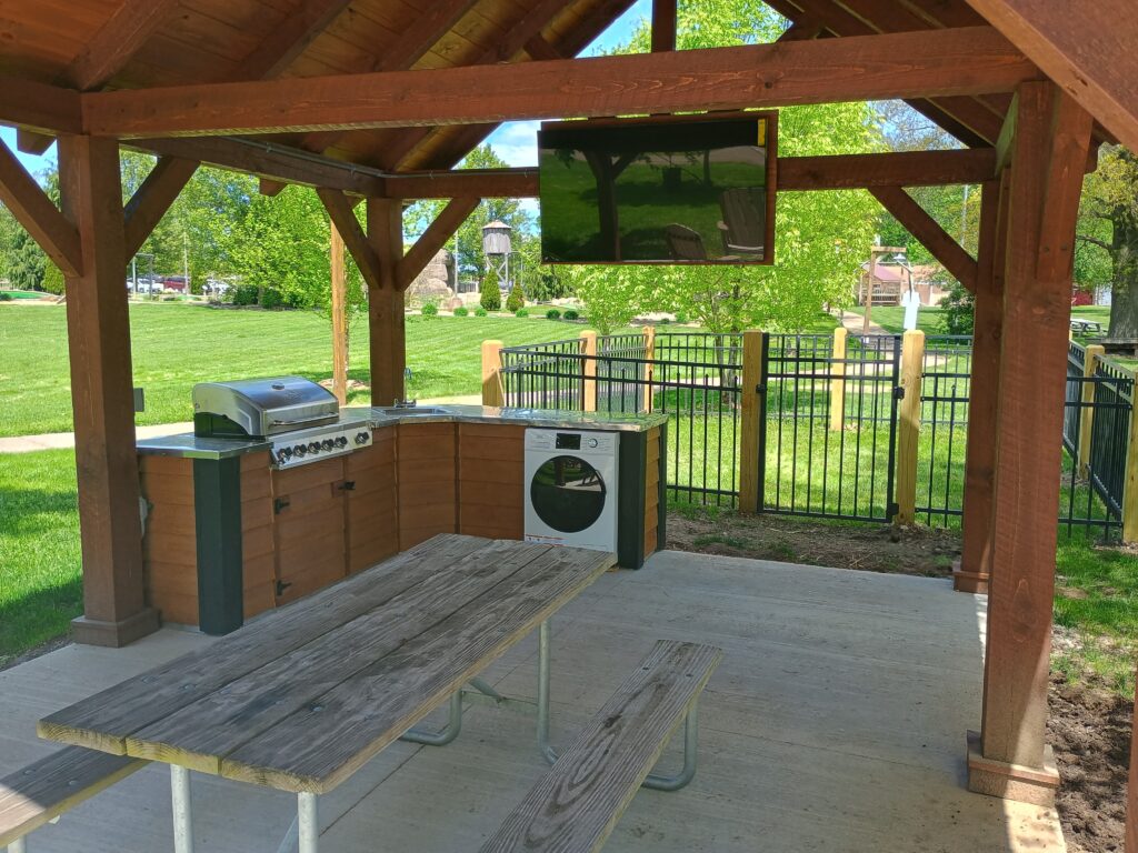 A covered outdoor kitchen with a grill, cabinets, a washing machine, a TV mounted overhead, and a wooden picnic table on a concrete patio in a park setting.