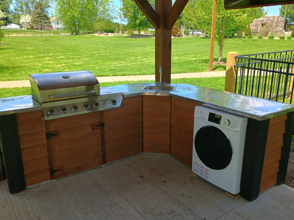Outdoor kitchen setup with a built-in grill, stainless steel countertop, sink, and a front-loading washing machine under a wooden shelter. Grass and trees are visible in the background.