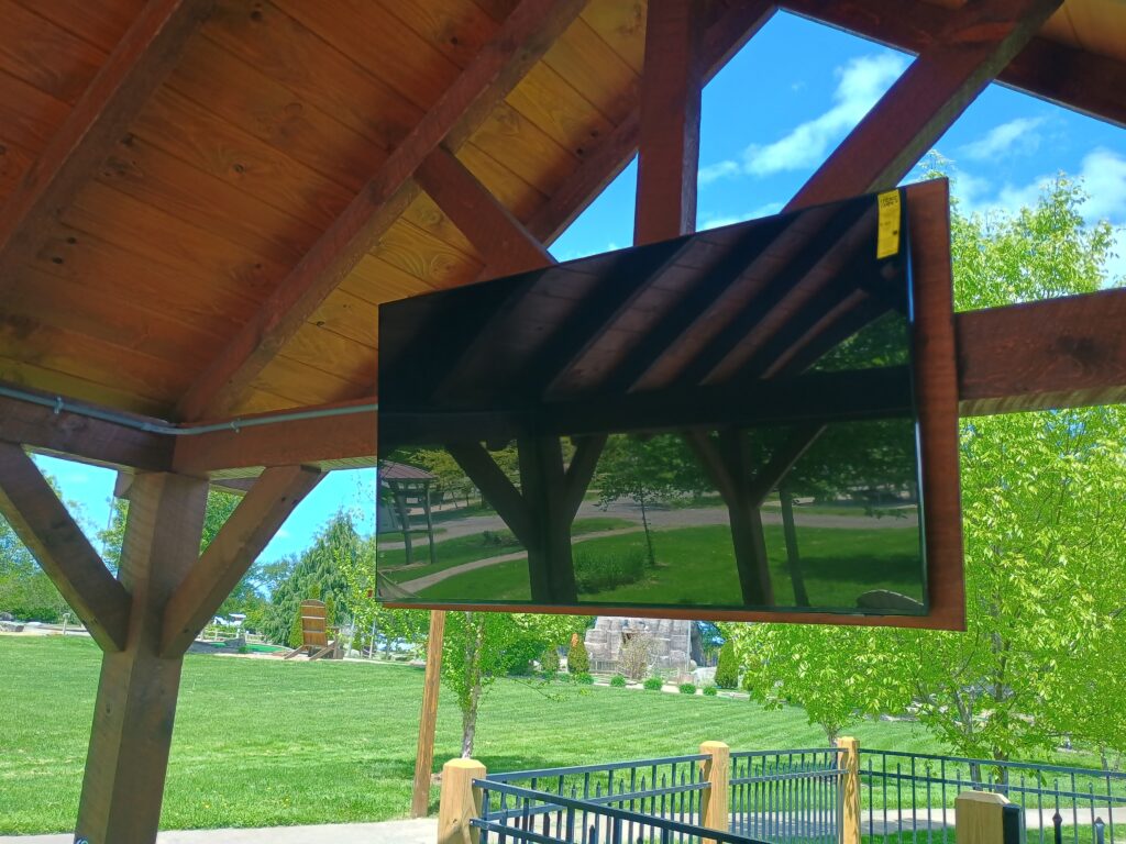 A flat-screen TV is mounted under a wooden pavilion, reflecting the beams, grassy lawn, trees, and blue sky in the background.