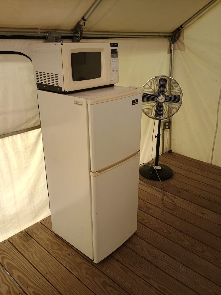 A microwave sits on top of a small refrigerator next to a standing fan inside a tent with wooden flooring, offering added comfort at campsites.