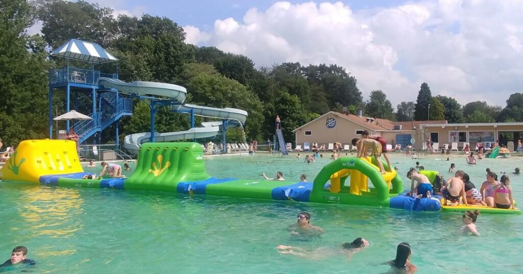 Outdoor pool with people swimming, playing on a floating inflatable obstacle course, and a large blue water slide in the background on a sunny day.