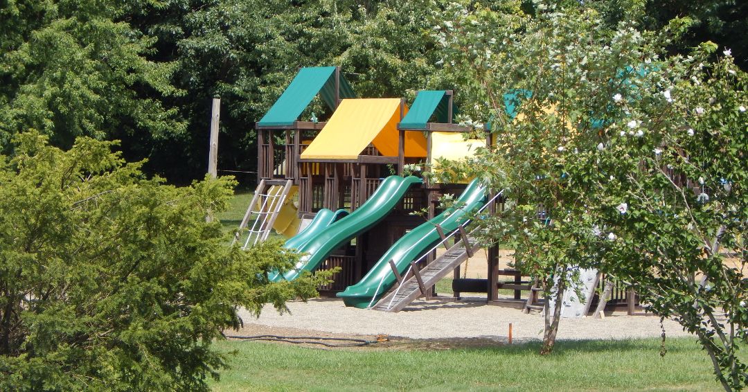 A playground with several green slides, yellow and green canopies, ladders, and climbing structures surrounded by trees and grass.