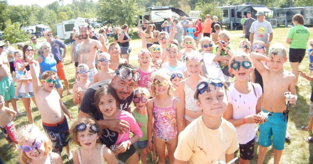 A large group of children and adults, many wearing swim goggles, smile at the camera during an outdoor gathering at a campground on a sunny day.