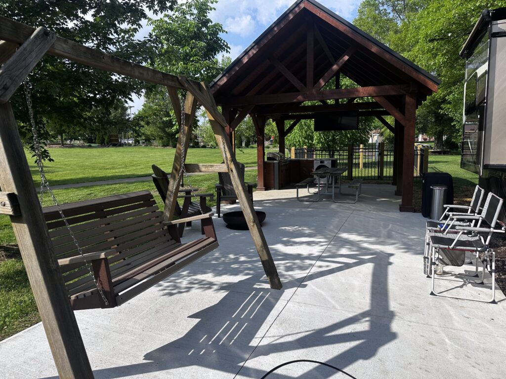 A covered outdoor patio at campsites with a wooden swing, chairs, a grill, a fire pit, and a TV, surrounded by trees and grass on a sunny day.