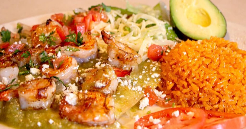 A plate of grilled shrimp over green sauce with rice, lettuce, sliced avocado, and diced tomatoes, garnished with crumbled cheese.