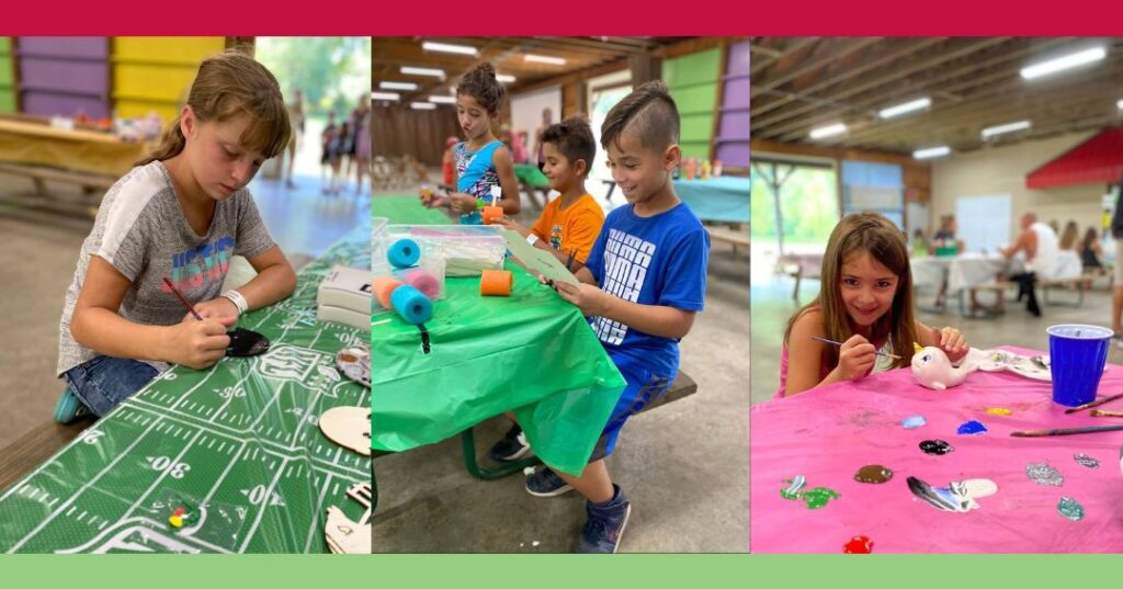 Children sit at tables indoors, painting various objects and crafts, with art supplies and colorful tablecloths spread out in front of them.