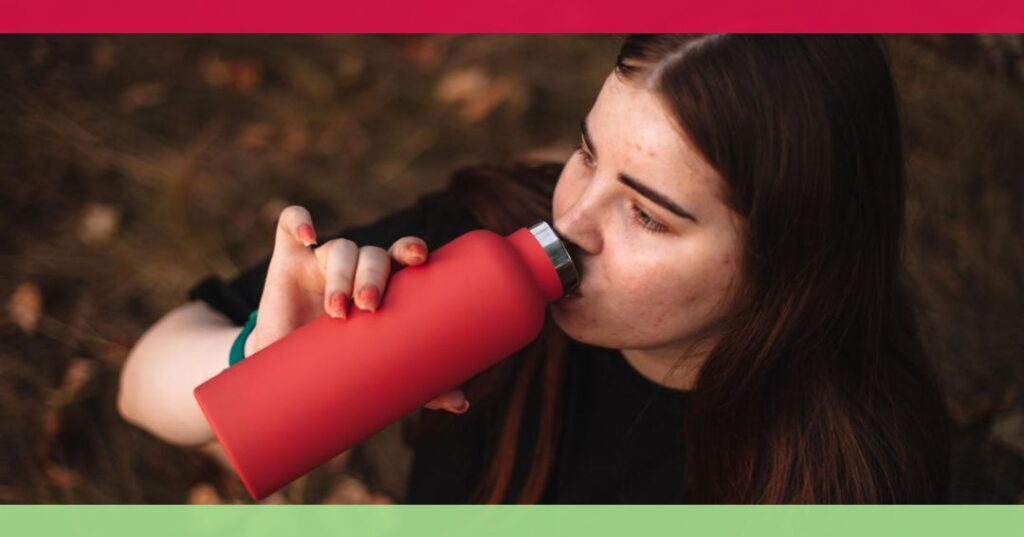 A young woman with long brown hair drinks from a red reusable water bottle outdoors.