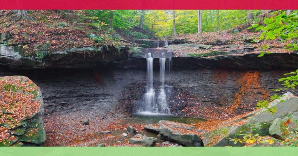 A small waterfall flows over a rocky ledge into a shallow pool, surrounded by autumn foliage and fallen leaves in a forest setting.