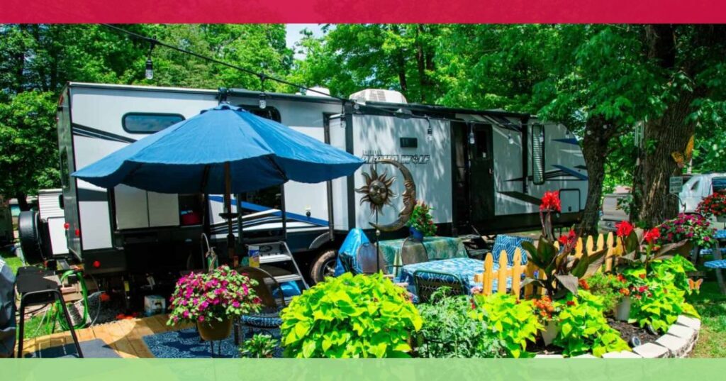 A travel trailer parked in a lush, green campsite with patio furniture, a blue umbrella, potted flowers, and decorative garden items in the foreground.