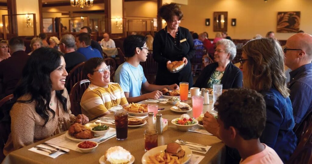 A group of people sit around a restaurant table enjoying a meal, while a server delivers a basket of bread. The table is set with food and drinks. The restaurant is busy with other diners.