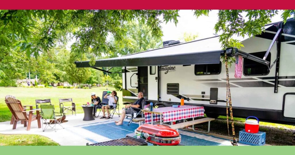 A family sits outside a parked RV under an awning, with chairs, a picnic table with a red checkered tablecloth, and various outdoor items on a sunny day.