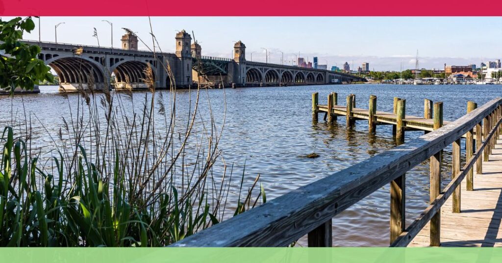 A riverside boardwalk leads to a wooden dock, with a large stone bridge and a city skyline visible in the background under a clear sky.