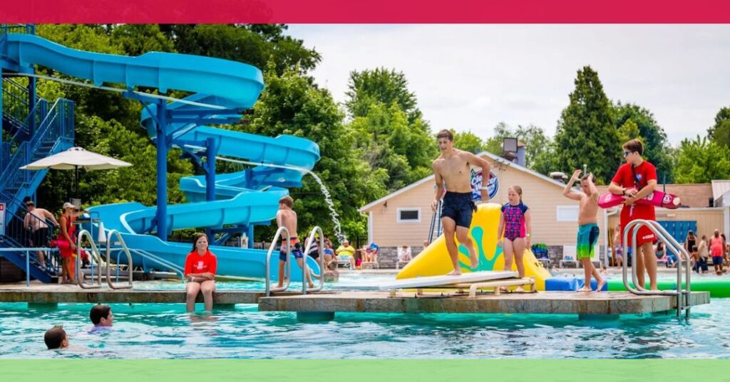 Children and lifeguards at an outdoor pool with a blue water slide, surrounded by trees and buildings on a sunny day.