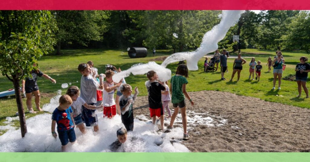 Children play in a large pile of soap foam outdoors while adults and other children watch in the background on a sunny day in a park.