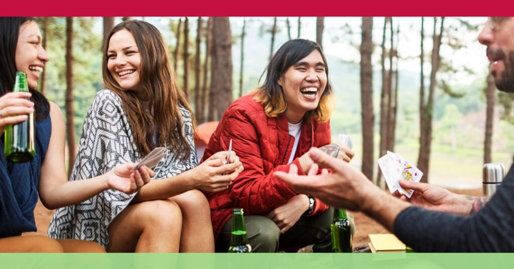 Four people sit outdoors in a wooded area, laughing and holding playing cards and drinks, with bottles on the table in front of them.