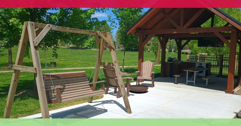 Wooden swing and Adirondack chairs on a concrete patio next to a covered pavilion with a picnic table, grill, and TV in a green park.