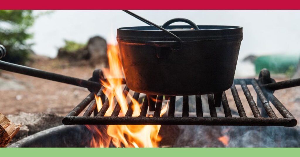 A black cast iron pot sits on a metal grill over an open campfire, with flames visible beneath, outdoors.