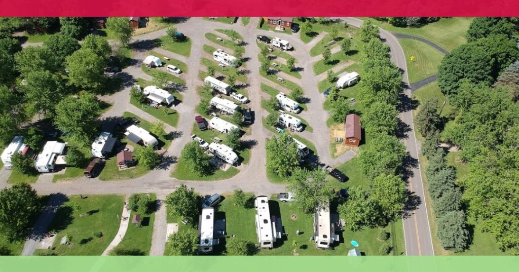 Aerial view of an RV park with multiple white RVs parked along curved gravel roads, surrounded by green trees and grass, with a paved road on the right side.