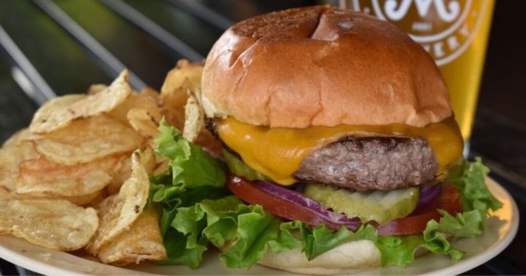 A cheeseburger with lettuce, tomato, pickles, and onion on a bun, served with potato chips on a white plate.