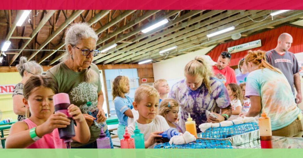 A group of children and adults gather at tables in a large indoor space, working on colorful crafts with bottles of dye and various art supplies.