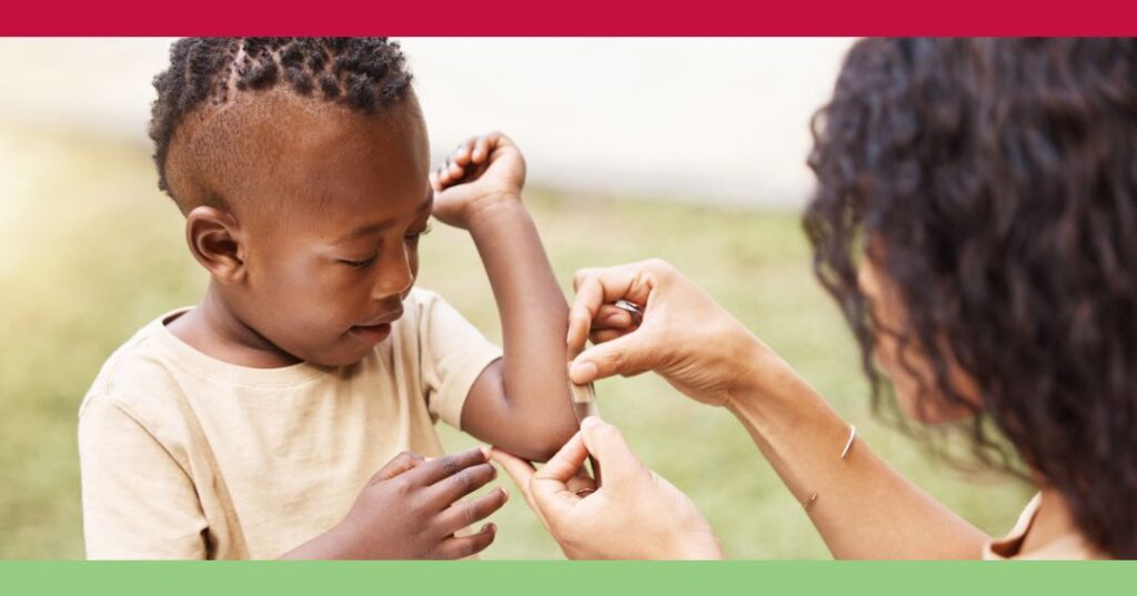 An adult applies a bandage to a young child's arm outdoors. The child lifts their sleeve while looking at the bandage.
