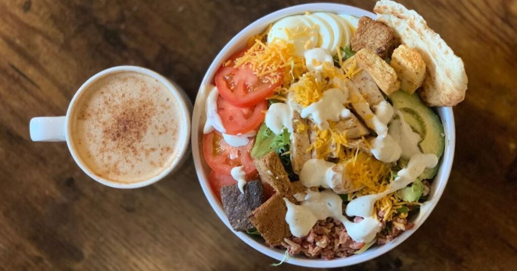 A bowl of salad with grilled chicken, sliced tomatoes, avocado, shredded cheese, egg, croutons, and ranch dressing beside a mug of coffee on a wooden table.