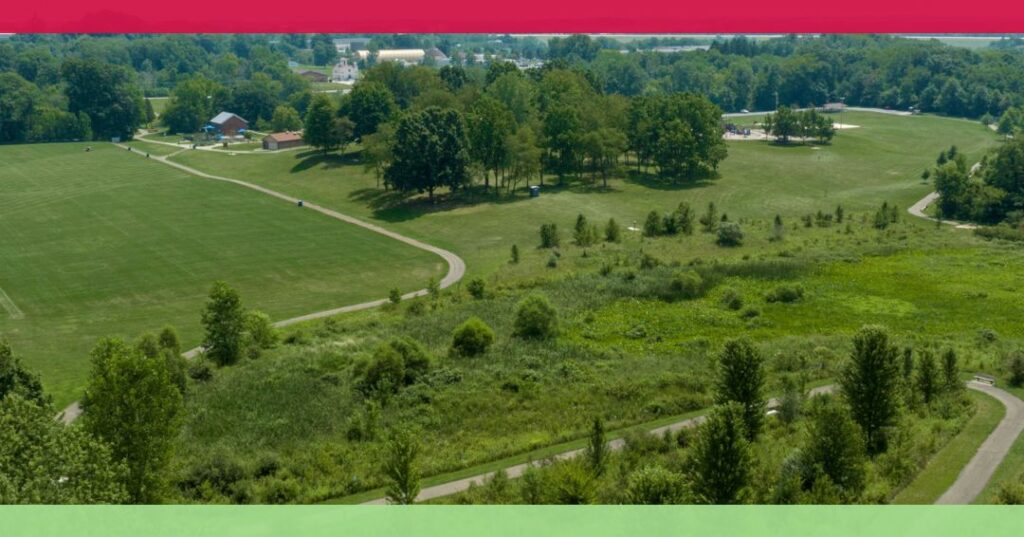 Aerial view of a park with open fields, walking paths, scattered trees, and surrounding greenery on a clear day.