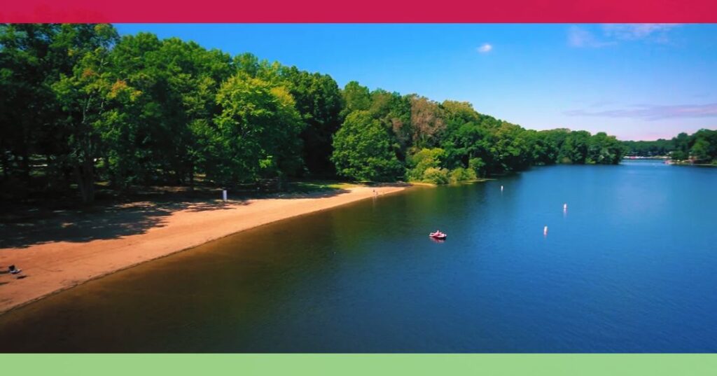 A sandy beach curves along a calm lake bordered by dense green trees under a clear blue sky. A person in a small boat is visible near the shoreline.