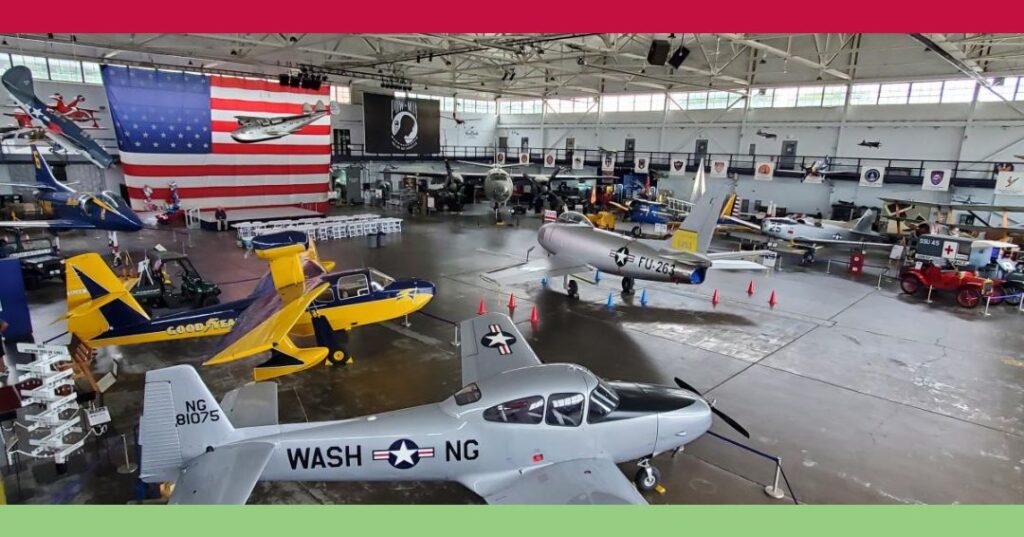 Aviation museum hangar with various vintage aircraft on display, large American flag in the background, and polished concrete floor.