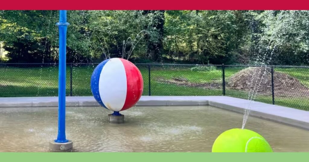 A splash pad with a large beach ball and a tennis ball sculpture spraying water, surrounded by trees and a black fence.