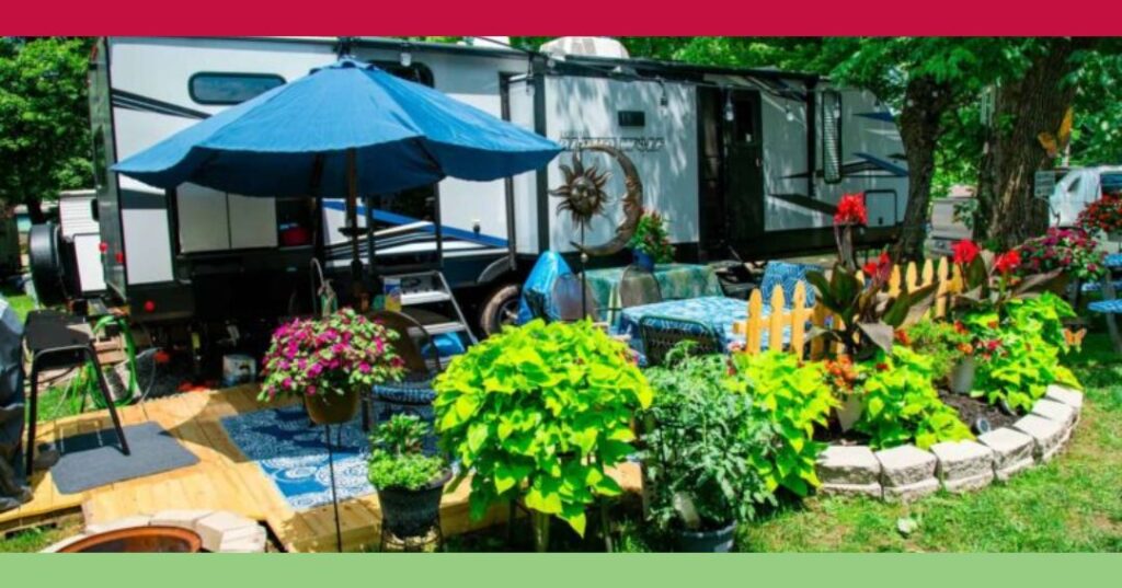A camper trailer with a wooden deck, outdoor furniture, an umbrella, and various potted plants and flowers arranged in a garden setting.
