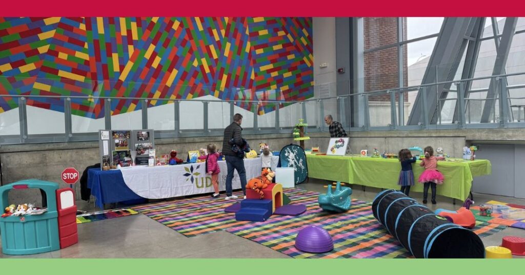 Children and adults interact at activity tables and play areas set up indoors on colorful rugs, with a bright multicolored mural on the wall in the background.